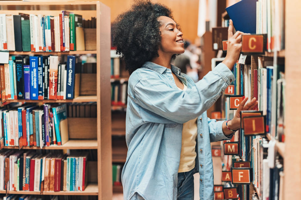 A woman is selecting a book from a library shelf, smiling. She's in casual attire