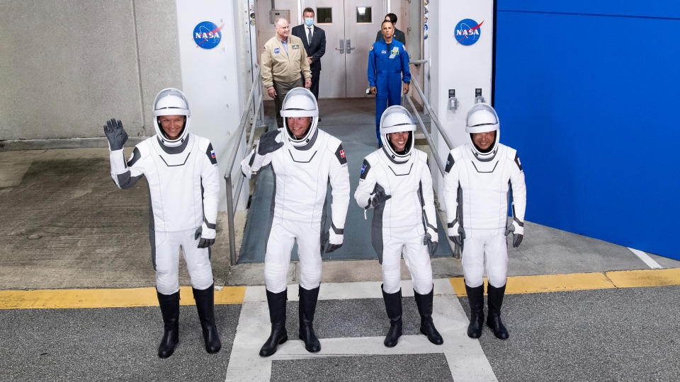 four astronauts in black and white spacesuits waving as they head to launch pad