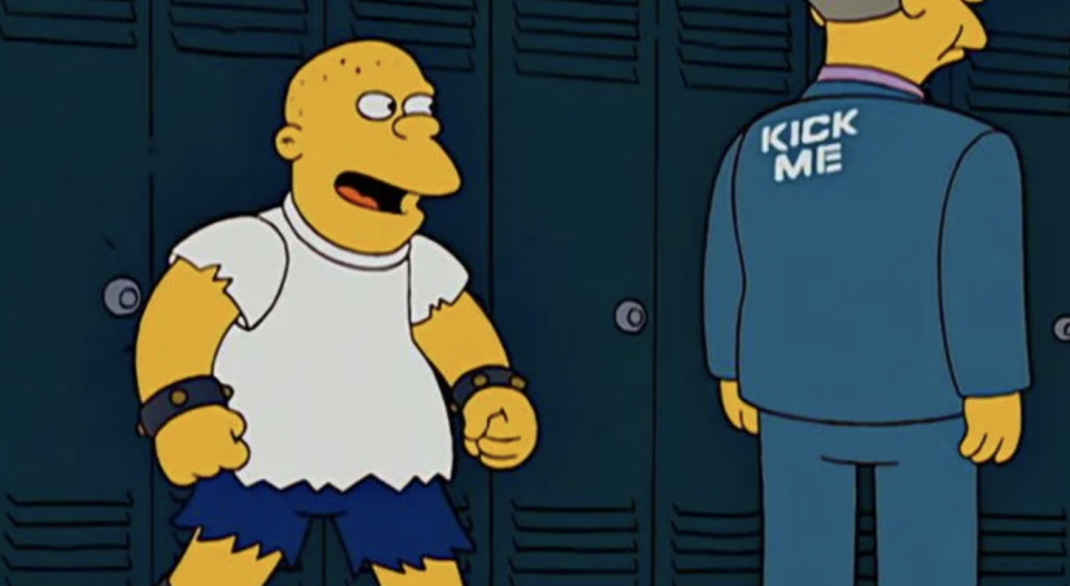 Kearney stands in a school hallway, laughing at Principal Skinner who has 'kick me' written on his back