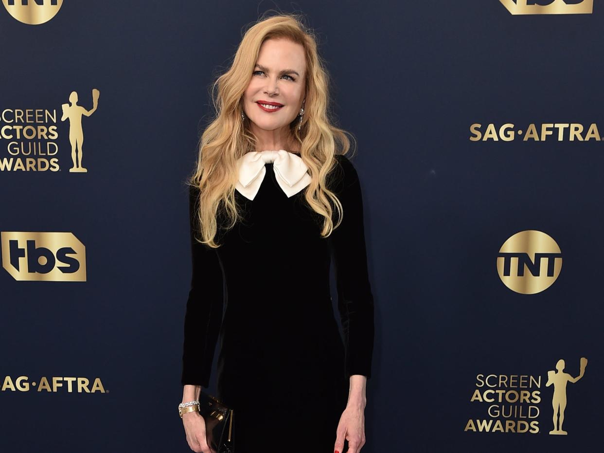 Nicole Kidman in a black dress with a white bow tie in front of an awards show backdrop.