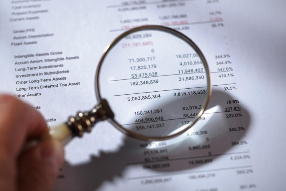 A hand holding a magnifying glass over a publicly traded company's balance sheet.