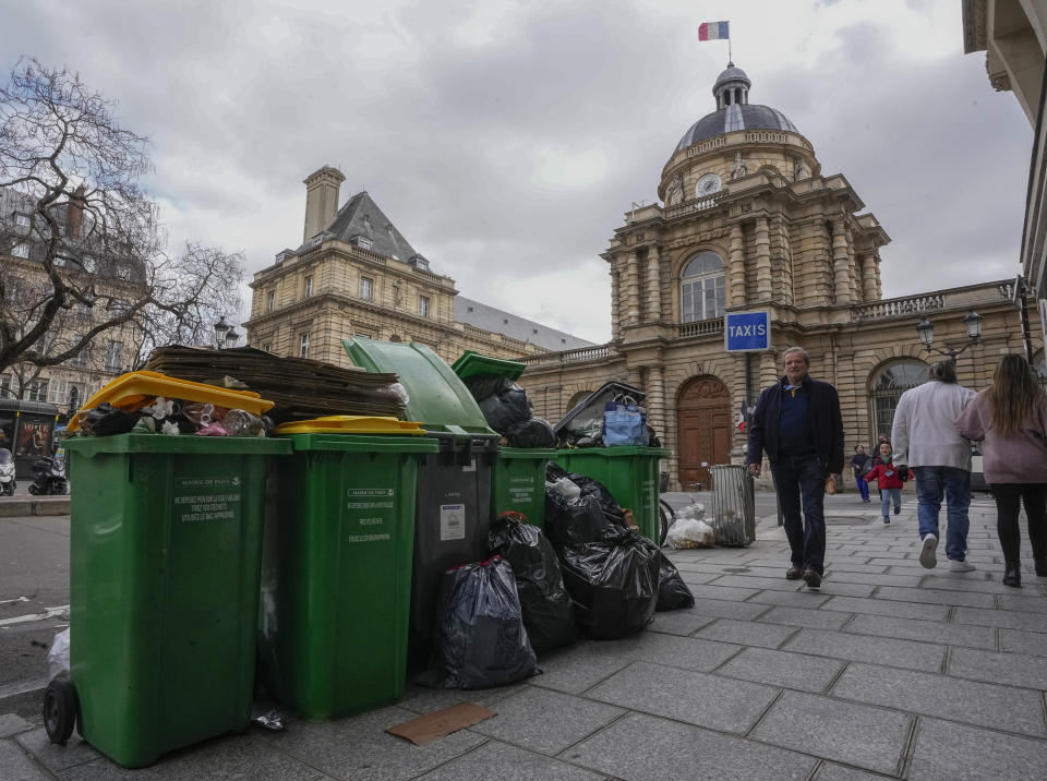 People walk past not collected garbage cans next to the Senate in Paris, Sunday, March 12, 2023. A contentious bill that would raise the retirement age in France from 62 to 64 got a push forward with the Senate's adoption of the measure amid strikes, protests and uncollected garbage piling higher by the day. (AP Photo/Michel Euler)