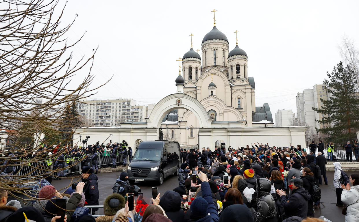 A hearse carrying Navalny’s coffin is parked outside the church before the funeral. Dozens of mourners stand behind barricades on either side of the vehicle.