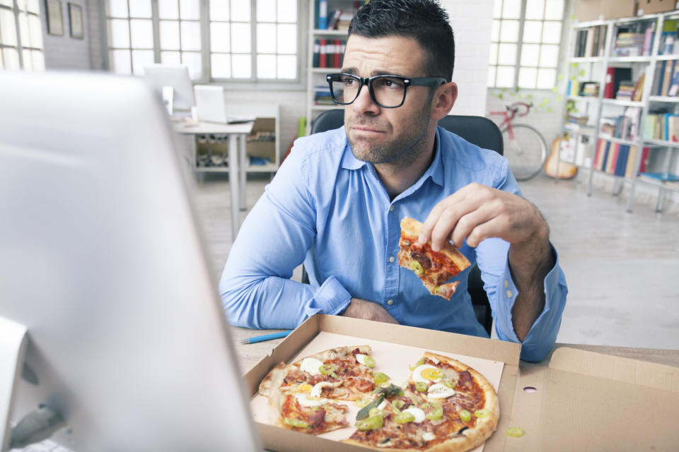 Tired young businessman holding slice of pizza and looking away. Man with eyeglasses and blue shirt. Monitor and pizza in box on desk. Windows, shelves with boxes and folders, desk with computer, bike and guitar as background.