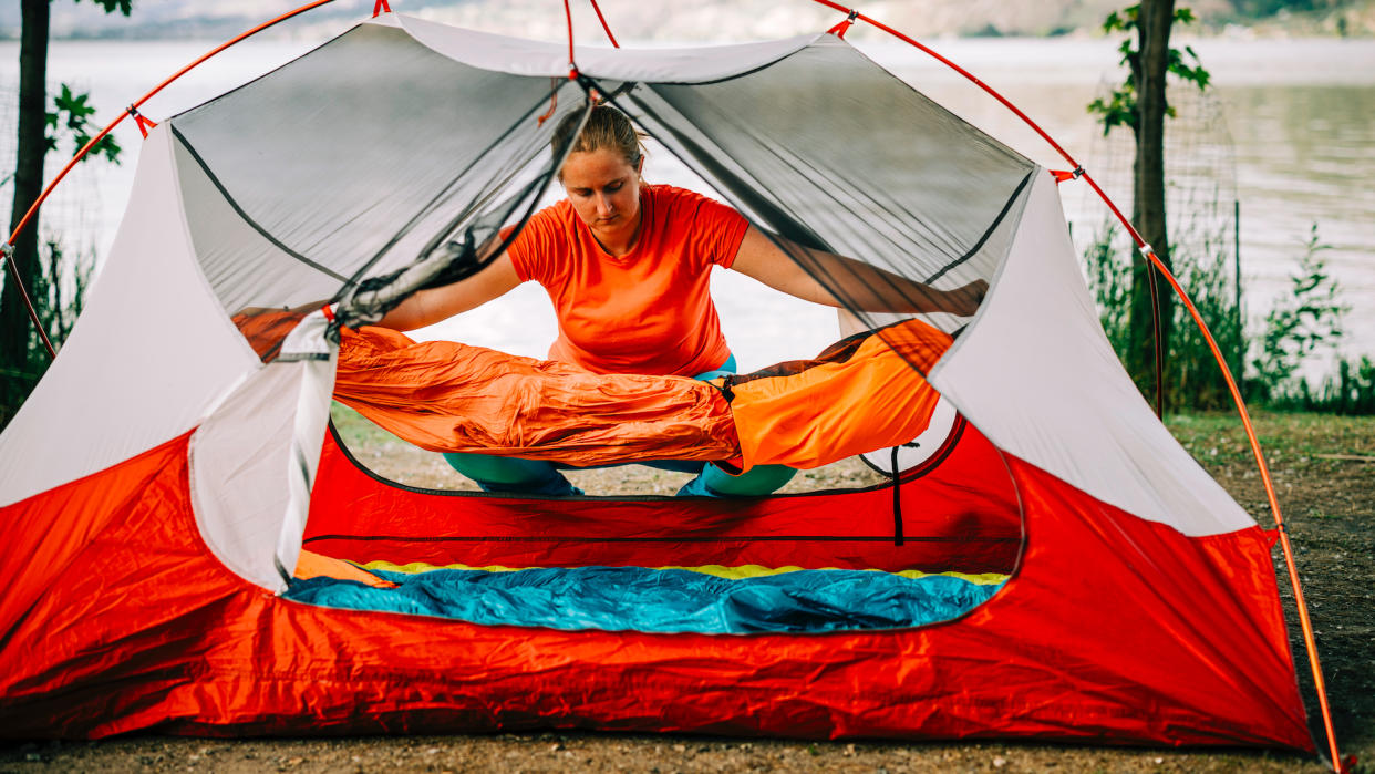  A woman lays out a sleeping bag inside a tent at a campground  – she’ll want a sleeping bag liner too. 