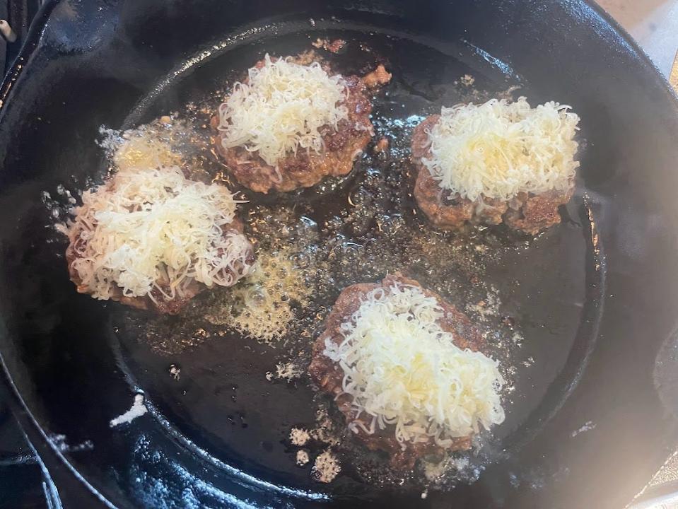 Four meat patties cook on a black skillet. Shredded Gruyere cheese melts on top of each patty