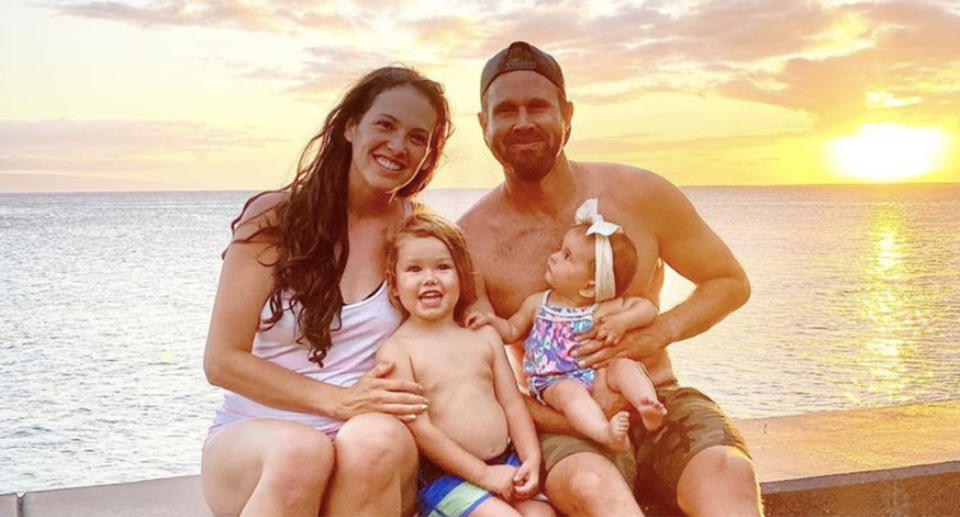 A photo of a woman and man smiling with a toddler and baby in front of the beach and beautiful sunset.