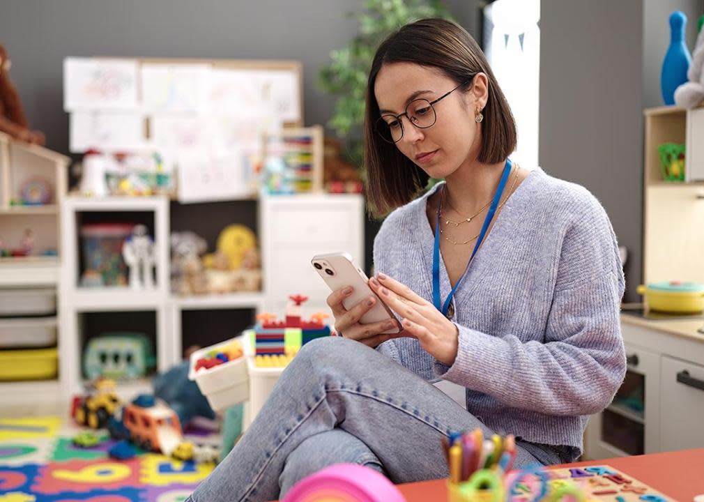 Young teacher sits in kindergarten classroom setting using smart phone