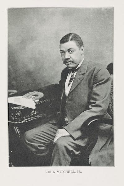 A Black man wearing a business suit sits at a desk with his right hand on a sheet of paper.