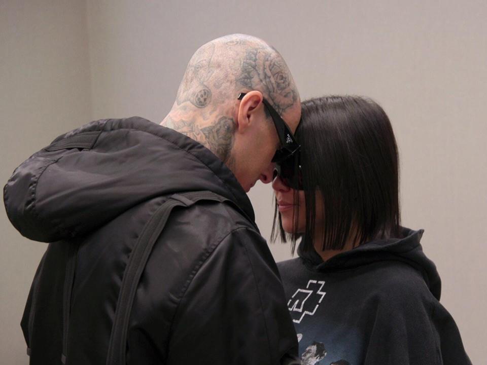 travis barker and kourtney kardashian solemnly touching foreheads. they're both wearing black hoodies and sunglasses