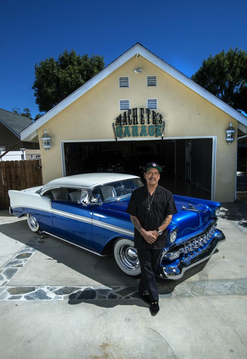 Danny Trejo in front of his 1956 blue and white car and a garage with the sign Machete's Garage