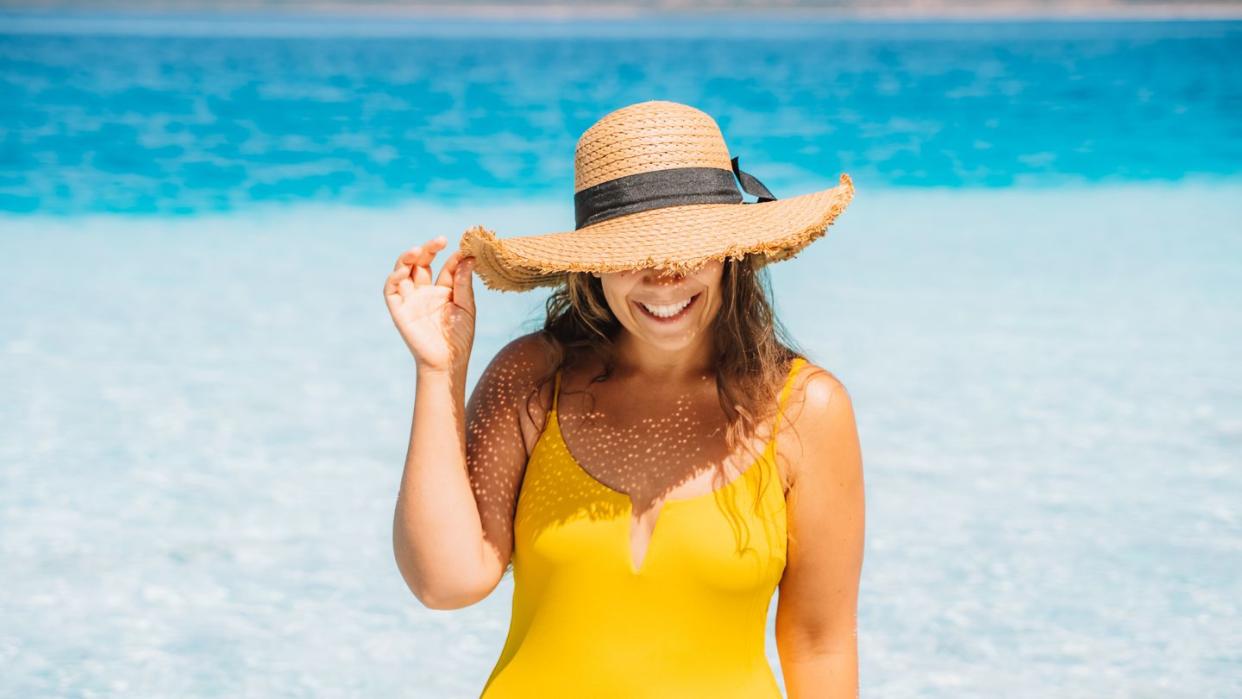 a woman in a yellow swimsuit and straw hat emerges from the sea with clear turquoise water on a summer sunny day