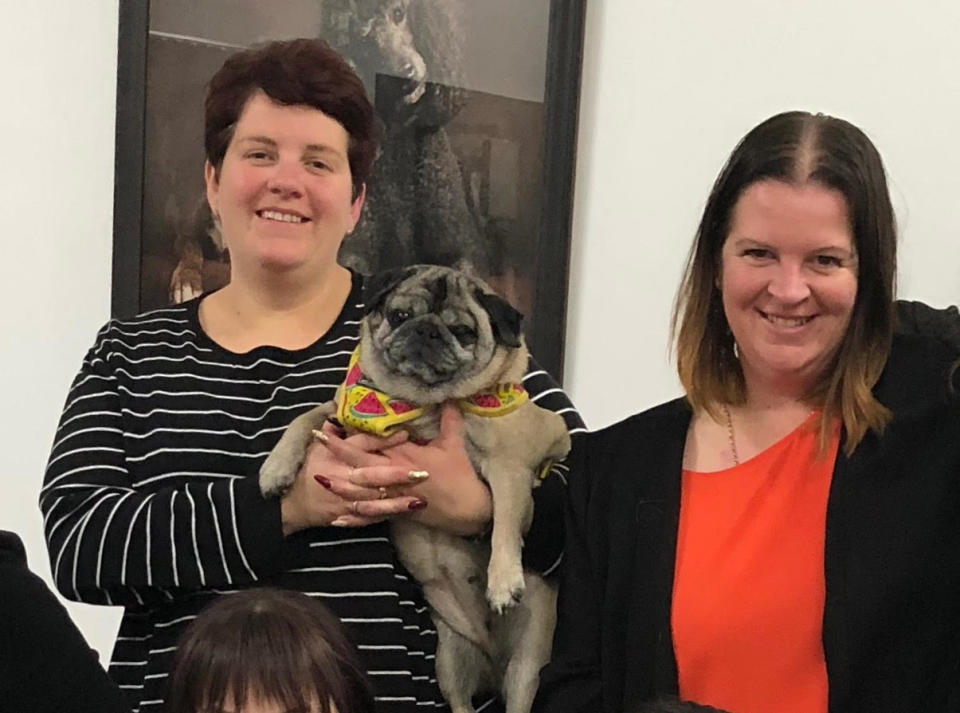 A woman wearing a navy and white striped top holds a pug dog next to a woman wearing a red top and black jacket.