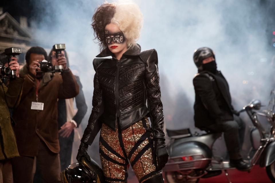 Cruella (Emma Stone) crashes a public event via motorcycle while donned in a glittering black leather jumpsuit with tire-tread shoulders.