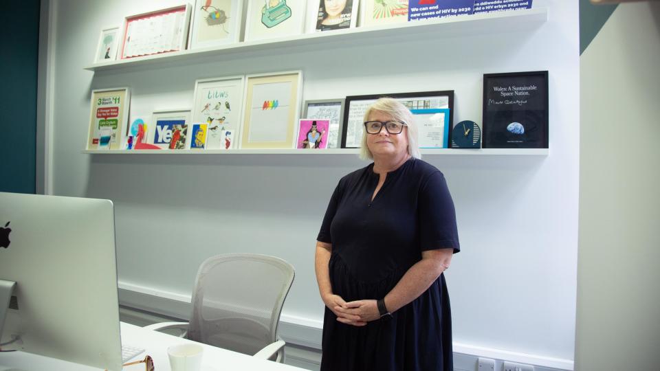 Woman with a black dress on and her arms across her stomach looking at the camera, there are frames of pictures behind her and an Apple monitor to the left