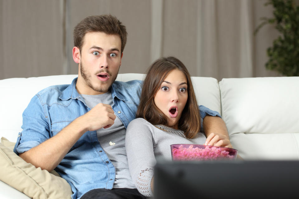 A young man and woman watching TV together, visibly shocked at what's on the screen.