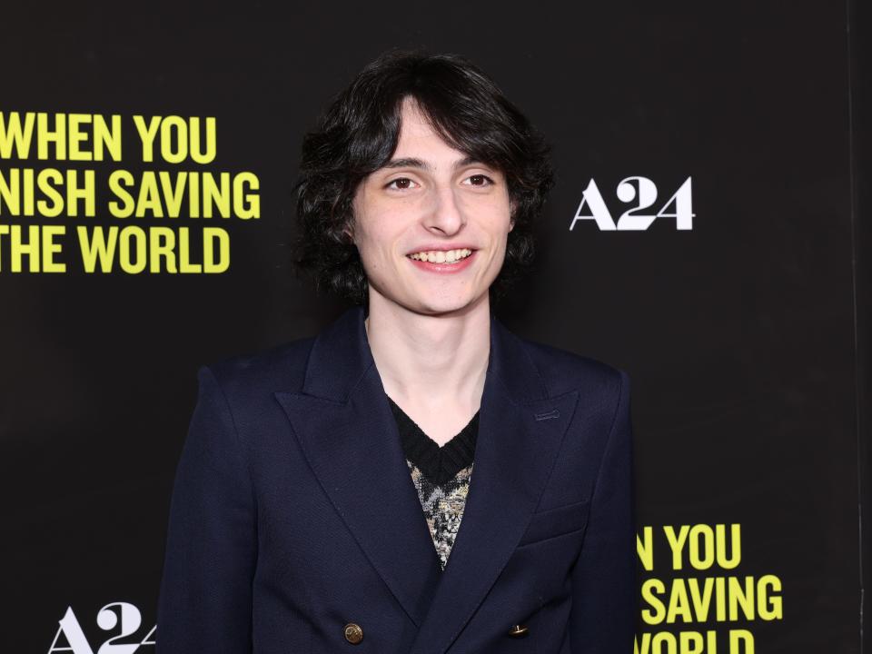 finn wolfhard at the when you finish saving the world premiere, standing on the red carpet. he's smiling, his hair styled around his face, adn wearing a buttoned navy blazer