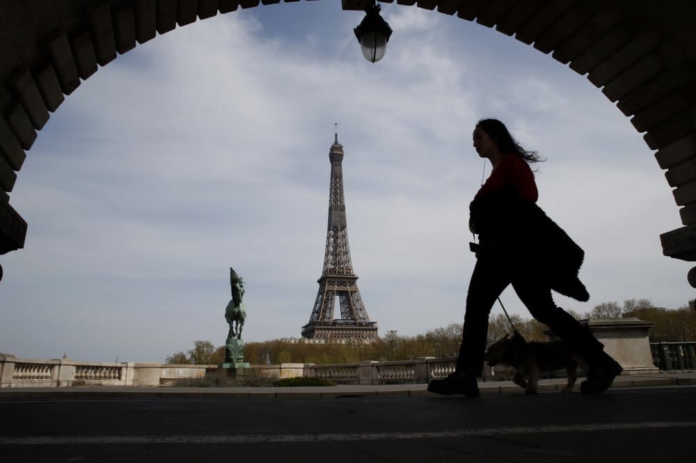 A woman walks her dog on a Paris bridge, with the Eiffel tower in background, during a nationwide confinement to counter the COVID-19. (AP Photo/Christophe Ena, File)