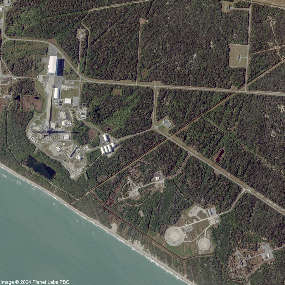 A view of Launch Complex 36 from space, February 14, 2024. an aerial view from space, looking down at a marshland adjacent to a pale sea. amongst teh overbrush wetlands, straights are cut out branching to different points. the leftmost with several developments, a large white building, and a concrete clearing. a long dark shadow stretches from the clearing center. below, diagonal along the shore, three smaller concrete pads are found through the branching straits.