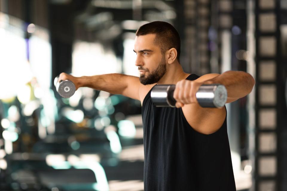 young muscular man doing dumbbell lateral raise exercise at gym