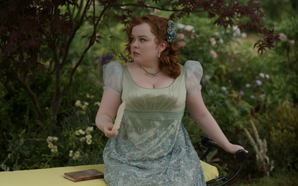 Penelope, in a floral lace gown with puffed sleeves, sits on a garden bench holding a letter