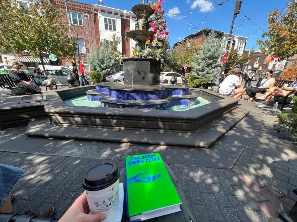 A woman holds a cup of takeout coffee and a book on her lap on a bench facing a fountain in the sunshine.