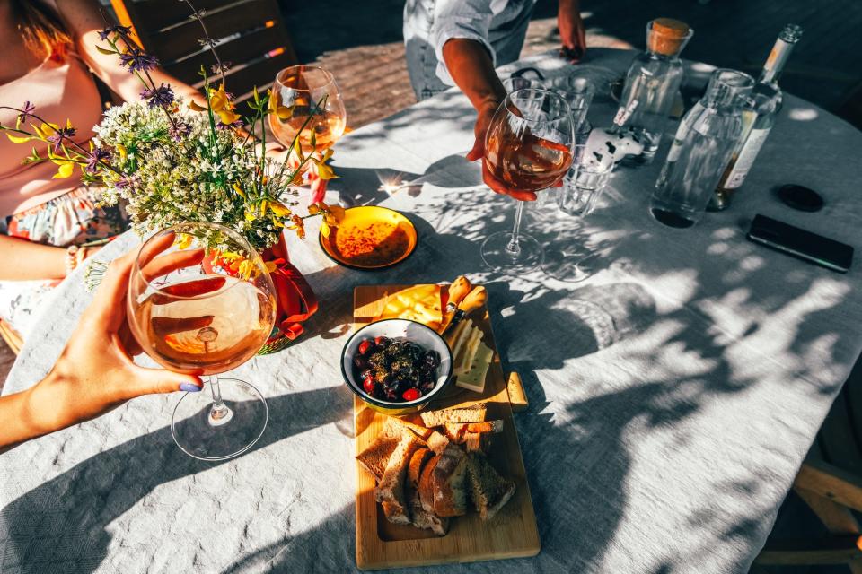 An image of an outdoor table with a plate of olives and bread, surrounding by people toasting with glasses of wine.