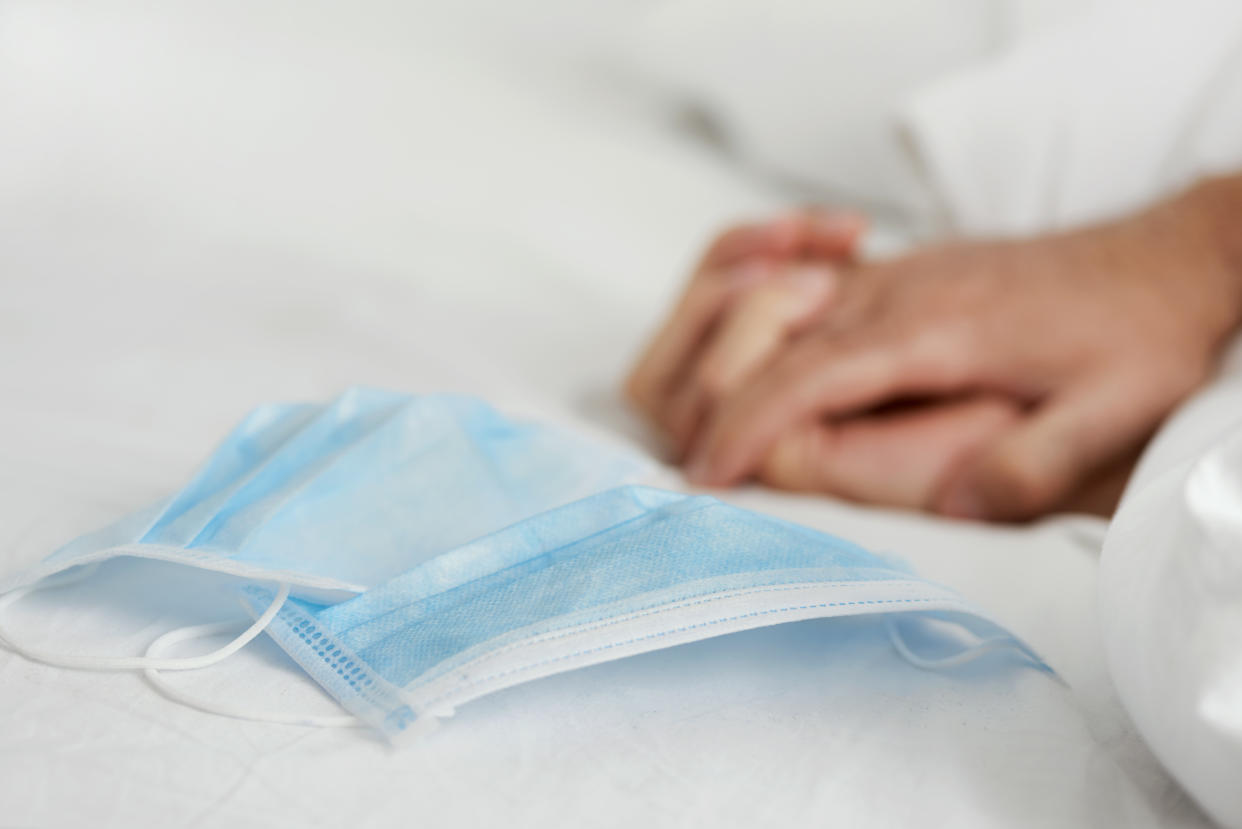 closeup of a pair of surgical masks on a bed set with white bedsheets, where two people are gripping their hands in the background