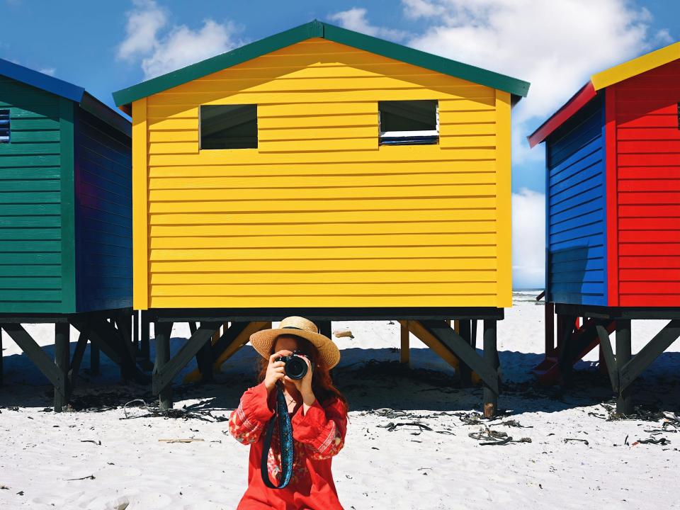 The writer kneeling in sand holding a camera in front of a small yellow house on a beach