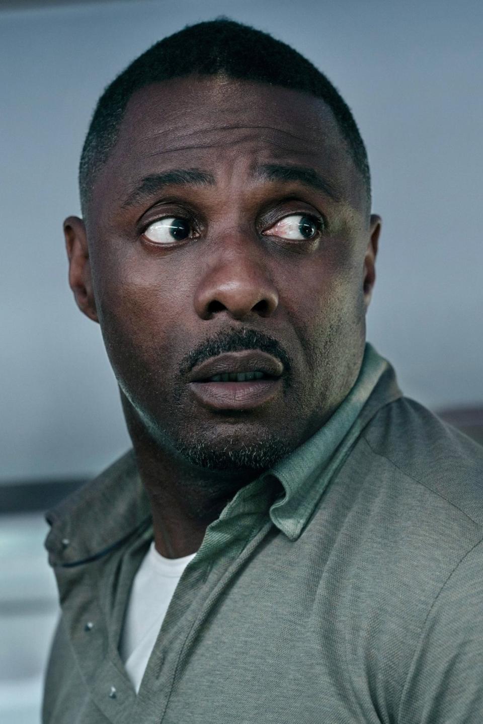 Idris Elba looking over his shoulder. He is wearing a buttoned-up shirt in a scene from a movie