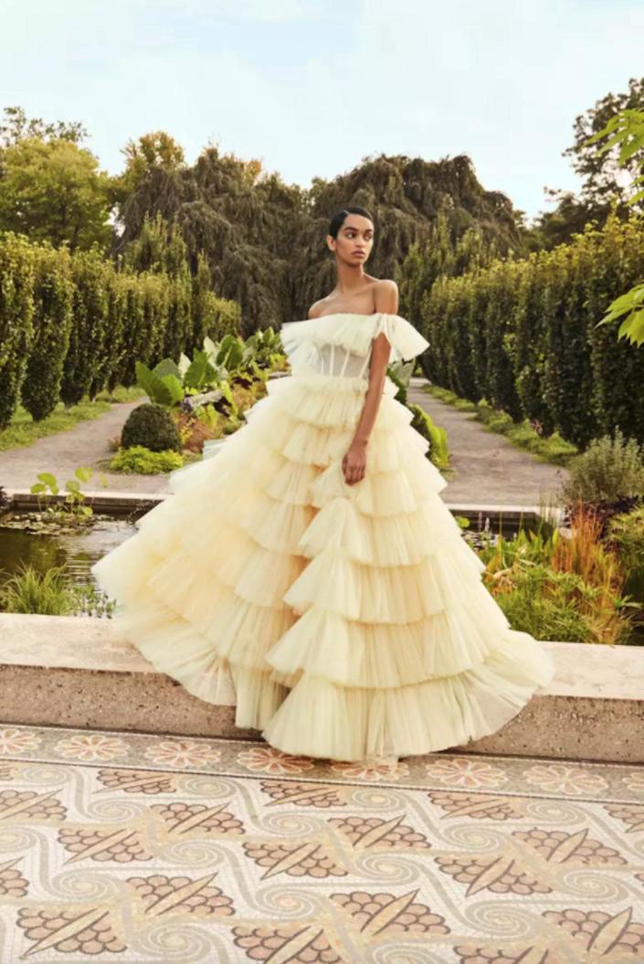 A bride poses in a garden wearing a yellow wedding dress with a corset bodice and a tiered skirt.