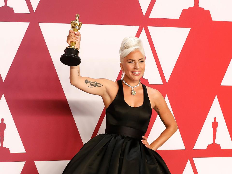 Gaga in a black tanktop-like ball gown holding her gold award in front of a red and white backdrop.