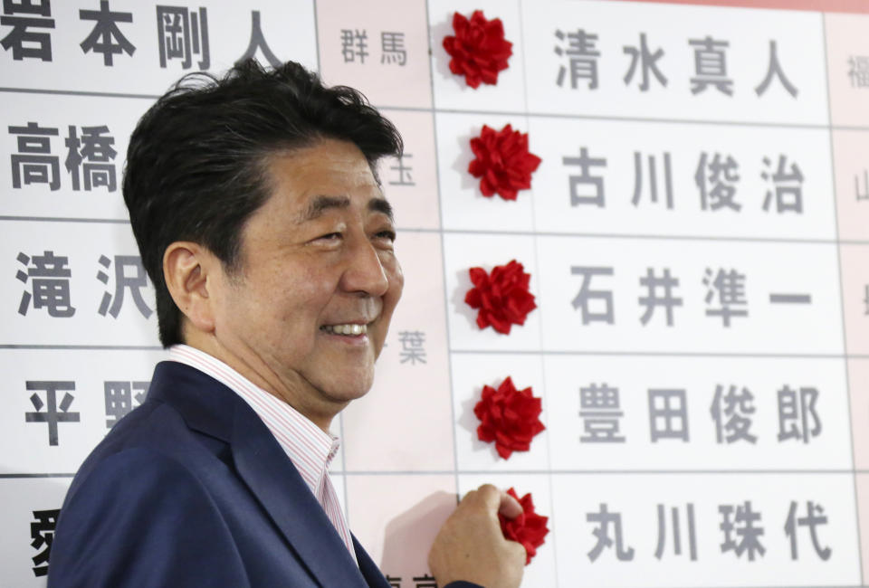 Japanese Prime Minister Shinzo Abe smiles in front of red rosettes on the names of his Liberal Democratic Party's winning candidates during ballot counting for the upper house elections at the party headquarters in Tokyo, Sunday, July 21, 2019. Prime Minister Abe's ruling coalition appeared certain to hold onto a majority in Japan's upper house of parliament, with exit polls from Sunday's election indicating he could even close in on the super-majority needed to propose constitutional revisions.(AP Photo/Koji Sasahara)