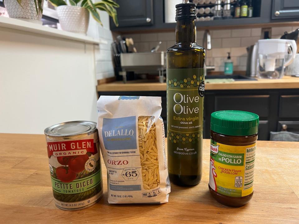 A wooden countertop with tinned tomatoes, a packet of orzo, a bottle of olive oil, and a tub of chicken bouillon.