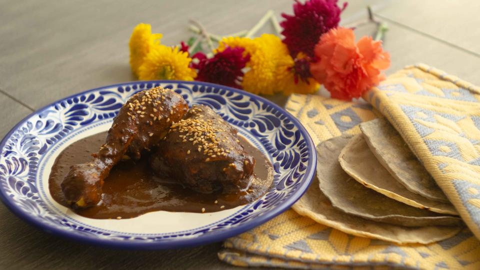 A wooden blue table with multi-colored flowers, corn tortillas inside a yellow cloth, and a blue and white Mexican plate with two pieces of chicken covered with Mexican mole sauce and sesame on top.