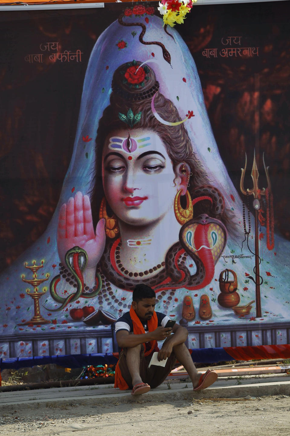 A Hindu pilgrim checks his mobile sitting next to an image of Hindu god Shiva at a base camp for the annual pilgrimage to the Amarnath cave shrine in Jammu, India, Monday, July 1, 2019. Thousands of Hindu pilgrims began the arduous trek to an icy Himalayan cave in disputed Kashmir on Monday, with tens of thousands of Indian government forces guarding roads and mountain passes. (AP Photo/Channi Anand)