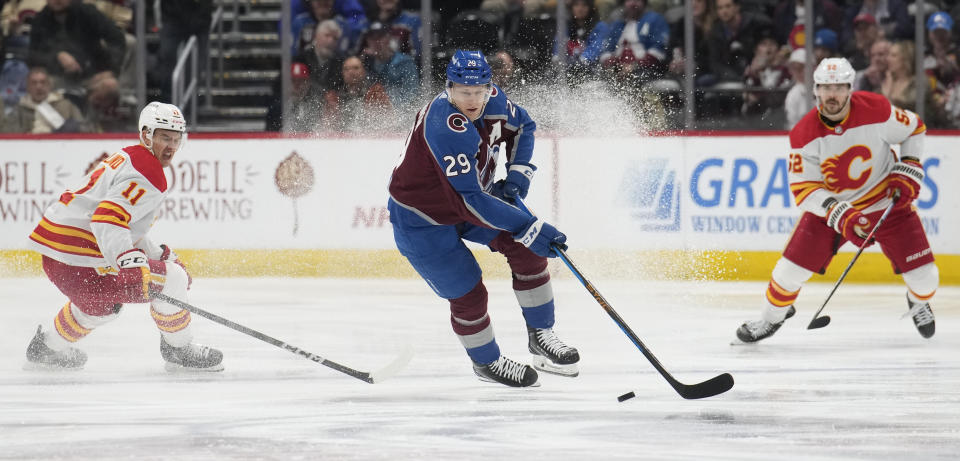 Colorado Avalanche center Nathan MacKinnon, center, picks up the puck between Calgary Flames center Mikael Backlund, left, and defenseman MacKenzie Weegar, right, in the second period of an NHL hockey game Monday, Dec. 11, 2023, in Denver. (AP Photo/David Zalubowski)