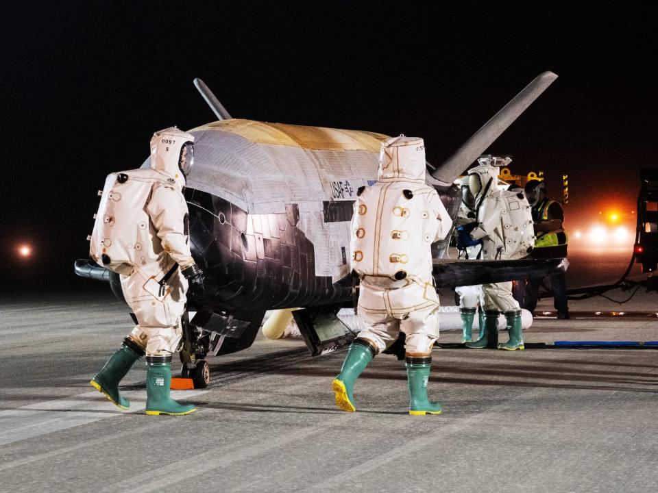 three people in puffy white protective suits approach a small white and black space plane on a runway