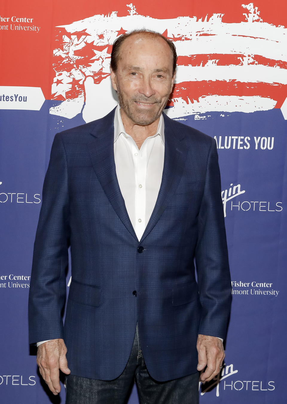 Lee Greenwood smiles at a red carpet event, wearing a white dress shirt under a dark blazer, while a banner behind him reads Fisher Center, Belmont University and Virgin Hotels.