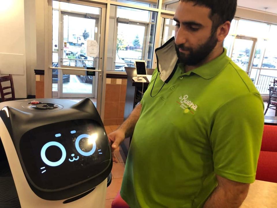 Elie Maalouf, owner of Gatineau, Que., brunch spot La Buena Déjeuner, poses with a cat-themed robot that now delivers meals to the restaurant's patrons.   (Hallie Cotnam/CBC - image credit)