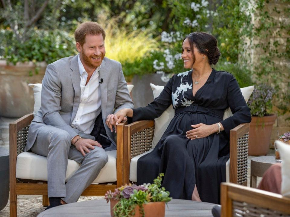 Harry and Meghan smiling and sitting outside and holding hands in formal attire.