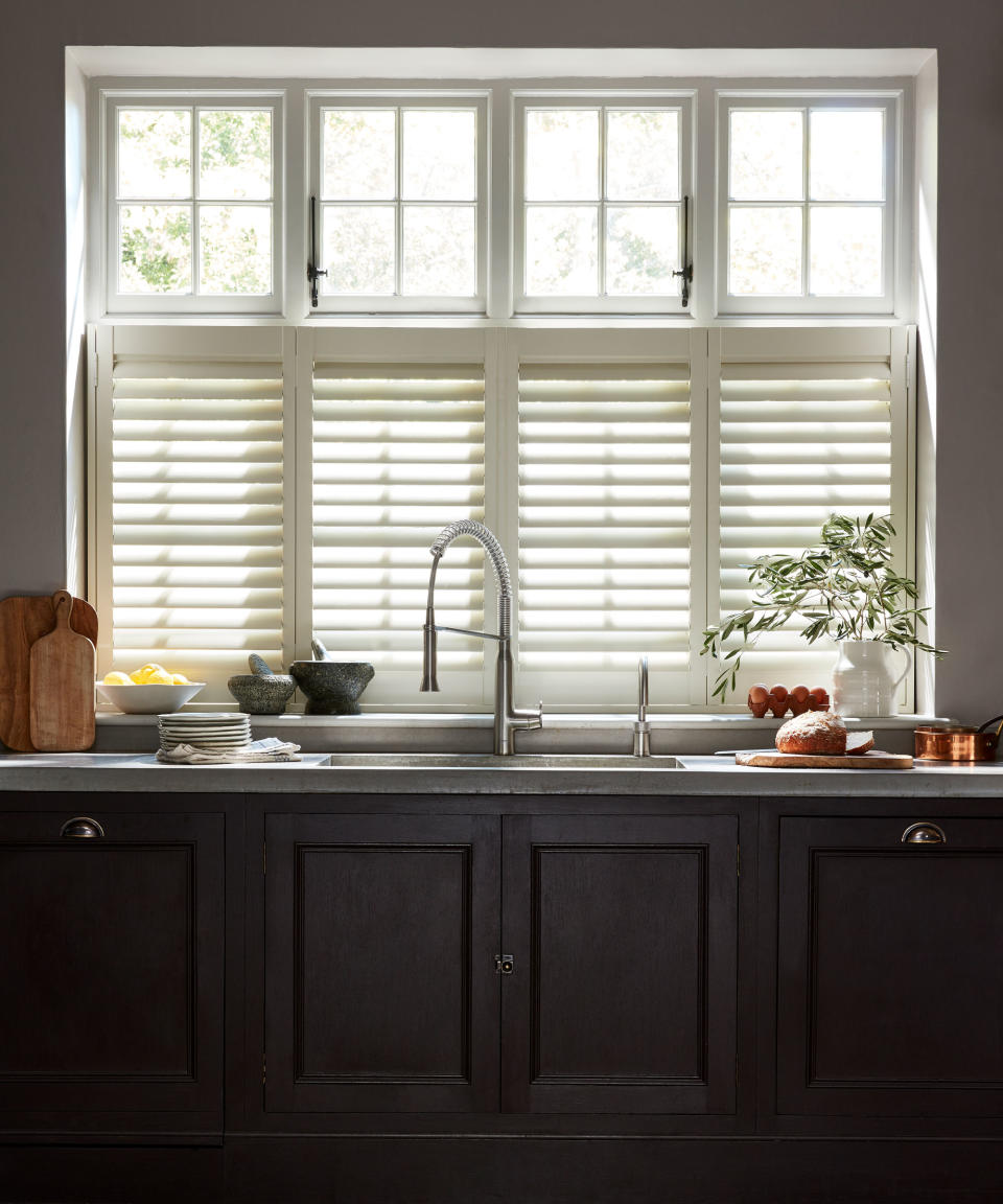 Large window above sink with white shutter, dark brown kitchen cabinets, kitchen sink, countertops decorated with kitchenware and food