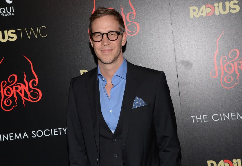 A man with short hair and glasses wearing a black suit and blue collared shirt