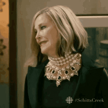 Moira Rose, a character from Schitt's Creek, is seen making a dramatic facial expression while wearing layered pearl necklaces and a dark outfit