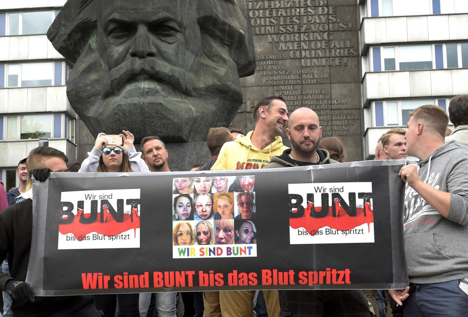 FILE - In this Aug. 27, 2018 file photo protesters gather for a far-right protest in front of a Karl Marx monument in Chemnitz, Germany. The large banner shows the bloodied faces of a dozen women above the words “we’re colorful until the blood flows.” The message to the boisterous crowd is clear: this is what migrants will do to you wives, sisters and daughters _ but the women pictured were actually victims of unrelated violent crimes in other countries. (AP Photo/Jens Meyer, file)