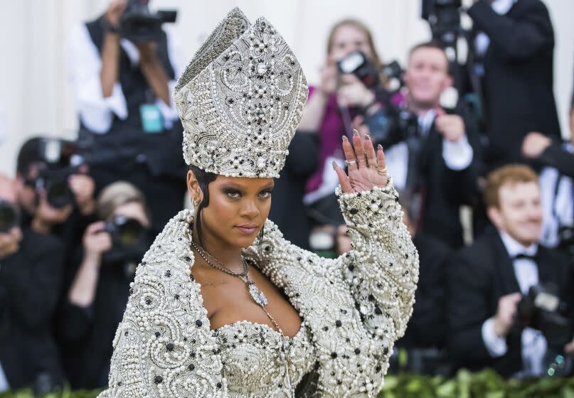 A woman waving while wearing a large bejeweled hat and matching coat, dress and necklace