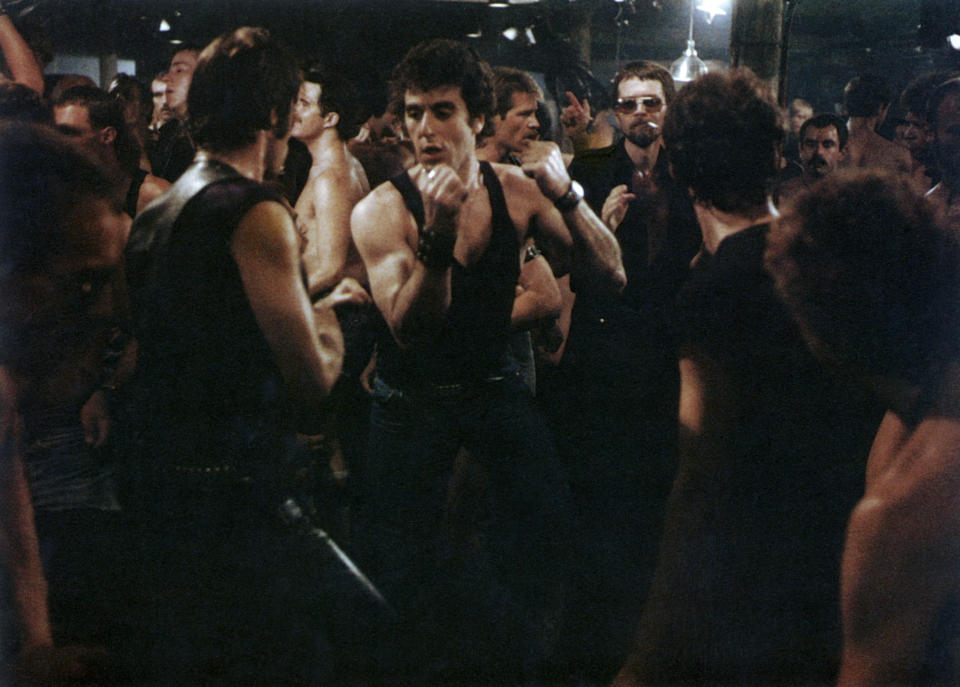 People in a crowded bar, most are shirtless or wearing sleeveless tops. A man in the center, flanked by others, appears to be dancing or fighting