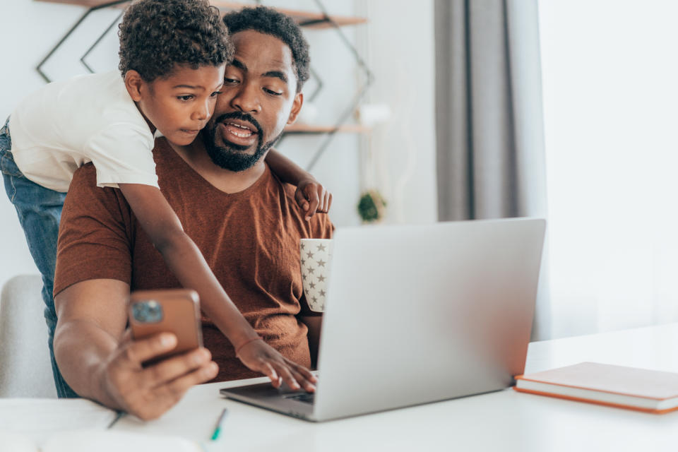 African American father using laptop and working at home while being distracted by his son.