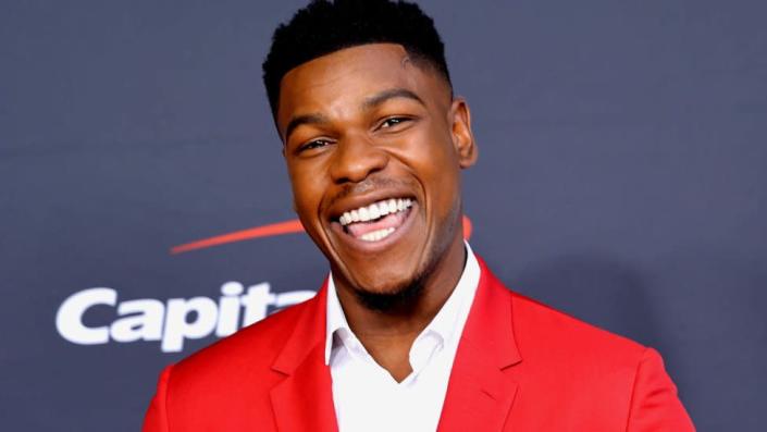 John Boyega in a bright red suit smiling widely.