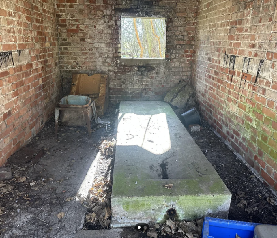 A dilapidated room with a decaying brick wall, a solitary chair, and remnants of a bed